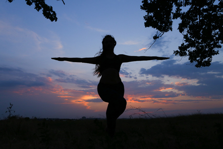 Female with beautiful figure takes yoga postures, gymnastics and ballet spends stretching and meditating, posing for advertising shoot fitness studio, healthy energy drink, dancing belly dancing among wide field on background of pink sunset and beautiful の写真素材