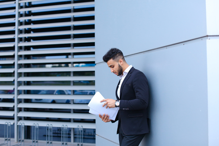 Perspective young adult businessman virile asian guy holds and looks, considers, reads and sorts out papers or documents, stands near gray wall of business center. Bearded man dressed in classic suit, black pants, jacket , white shirt, on hand is watch. Cの写真素材
