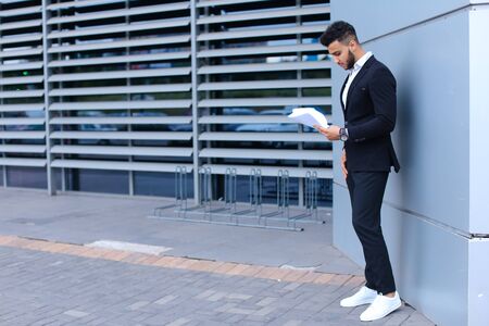 Young tall handsome arab entrepreneur guy student holds and considers, reads paper or documents, stands in full growth in profile near gray wall of business center. Bearded man dressed in classic suit, black pants, jacket , white shirt and sneakers,  on hの写真素材