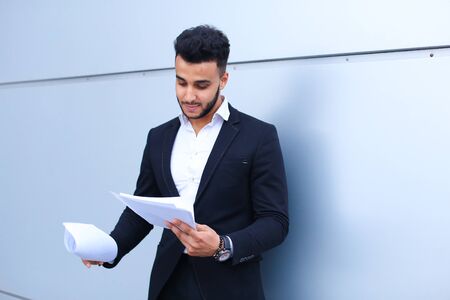 Young successful businessman arab man holds documents, securities, smiling little smile and thinks near gray wall of business center. Muscular bearded man dressed in classic suit, black pants, jacket , white shirt. Concept of securities, Business Meeting,の写真素材
