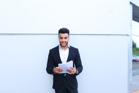 Young successful student businessman arab man holds documents, securities, smiling and thinks near gray wall of business center. Muscular bearded man dressed in classic suit, black pants, jacket , white shirt, on hand is watch. Concept of securities, Busiの写真素材