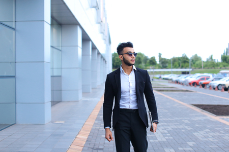 Young handsome businessman Arab Muslims is in the business center in black elegant dress on building background. Businessman man male looking at time walking goes and smiling.の写真素材