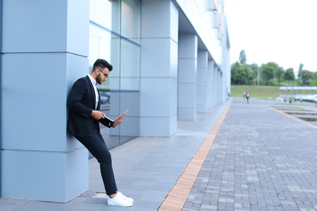 Entrepreneur uses  laptop wearing sunglasses. Young handsome businessman arab muslims in business center wearing dressed in black elegant suit on building background.の写真素材