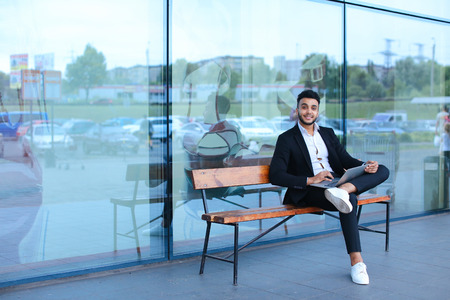Entrepreneur uses laptop wearing sunglasses. Young handsome businessman arab muslims in business center wearing dressed in black elegant suit on building background.の写真素材