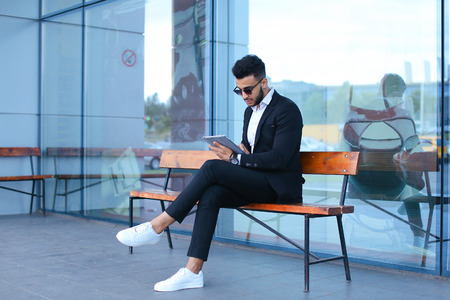 Entrepreneur smiling man  puts on sunglasses uses tablet and looks at. Young handsome businessman arab muslims in business center wearing dressed in black elegant suit on building background.の写真素材