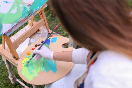 Pretty attractive artist begins to painting which depicts  colorful butterfly, sitting with back to photographer and behind wooden easel, holds palette smeared in paint in large park green day outdoors.Girl with brown hair collected in beam dressed in ligの写真素材