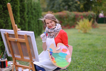 Cute female painter posing, laughing and in front of camera , draws and smiling with brushes and colorful palette in hand, sits on chair near easel in green park outdoor.  Girl with brown hair dressed in red jamper, dark denim overalls and white apron, chの写真素材