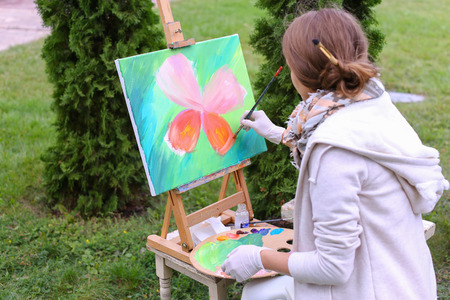 Pretty attractive artist finishes painting which depicts colorful butterfly and sitting with back to photographer, and behind wooden easel, holds palette smeared in paint in large park day outdoors.Girl with brown hair collected in beam dressed in light jの写真素材