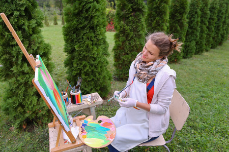 Female artist holds brush and wrote oil on canvas painting, does what likes, interests and hobbies,  preparing picture to order, advertises or art studio for drawing utensils, sits on chair at  wooden easel in large green park outdoors. Girl with brown haの写真素材