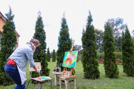 Young girl European appearance looks at drawing of Multicolored butterflies, which painted with oil paints and estimates finalizing remaining details. Stands near wooden easel with brush in hands on background art materials and coniferous trees in park ouの写真素材