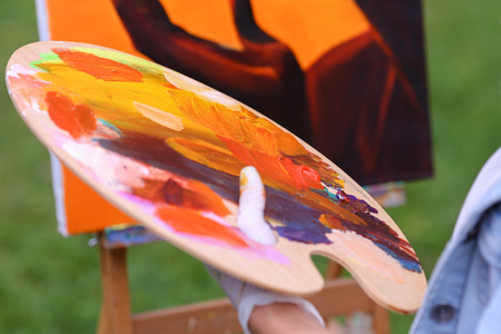 Photo of female hand artist who mixes oil or acrylic colored bright colors on wooden panel, which would start to draw and located in green park in open air. Concept of set of objects to paint, oil or acrylic paints, advertising art school.の写真素材