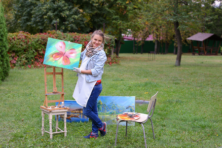 Female European appearance artist evaluates picture and removes from easel, holds and posing with picture which depicts colorful butterfly, smiling and standing in full growth on background of lawn and angered by conifers in large park in open air. Girl dの写真素材