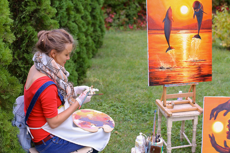 Artist cleans workplace, evaluate work that stands on easel, smiling, posing and looking in camera, sits on chair near paintings in park outdoors on background of coniferous plants in afternoon. Girl dressed in red sweater, dark jeans, white apron and cheの写真素材