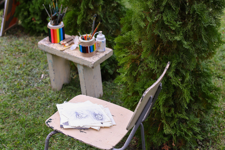 Photo which shows chair with pencil sketches, which stands near gray stool on which stand with colorful jars with thick and thin brushes for oil and acrylic paints and colored pencils. Photo taken in park on grass background in daylight.Concept of art matの写真素材