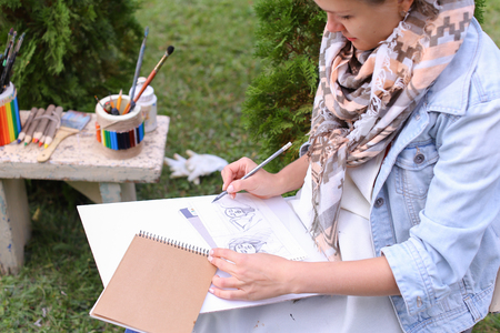 Contemporary Artist Works on Facial Contour of Depicted on Portrait Unknown Girl, Draws Sketch of Fictional Head, Prepares Gift For Friend. Woman Located in Park Outdoors in Background Stands Bench With Colors and Additional Art Materials. Artist Dressed の写真素材