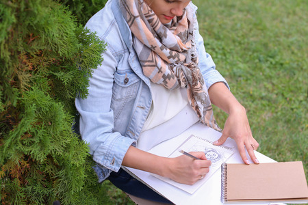 Contemporary Artist Works on Facial Contour of Depicted on Portrait Unknown Girl, Draws Sketch of Fictional Head, Prepares Gift For Friend. Woman Located in Park Outdoors in Background Stands Bench With Colors and Additional Art Materials. Artist Dressed の写真素材