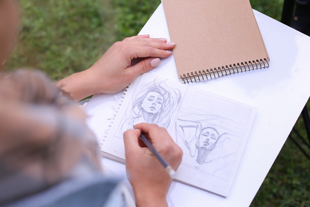 Talented Young Woman Handles Details ,obscures Hair in Picture. Sketch Executed Ordinary Pencil on Sheet of Paper Graphical Way, Which Lying on Wooden Tablet on Artist Knees, Which Located in Park in Open Air. Artist Dressed in White Jacket, Jeans and Cheの写真素材