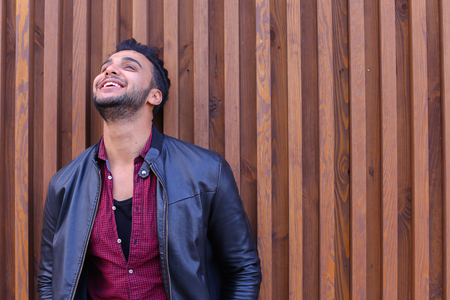 Engaging Young Adult Arabian Male Man, Businessman, Student Smiles and Laughs, Posing For Camera and Winks, Looks Forward on Sides, Waits Meeting, Corrects Hairstyle and Recalls Something Good Stands Near Wood-Paneled Walls in Open Air. Confident Guy Arabの写真素材