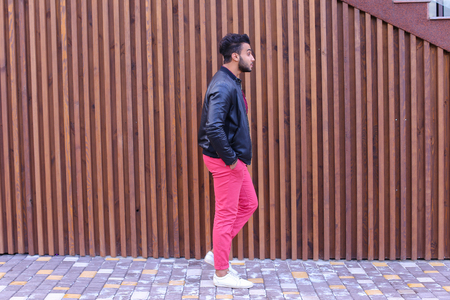 Handsome Young Adult Muslim Man, Businessman Walks Slowly Along Stairs, Looks Around and Smiles, Holds Hands in Pockets of Jackets, Advertises Clothing on Background of Wooden Stairs Near Restaurant Outdoors. Confident Arab Guy With Dark Hair Dressed in Bの写真素材