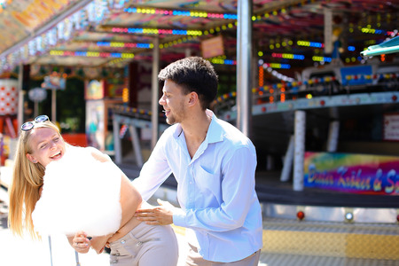 Beautiful young people, loving couple strolling in amusement park, looking at each other, kissing and cuddling, try candy floss, pose for camera and fooled at an amusement park outdoors. Girl with long blond hair and sunglasses dressed in white top and beの写真素材