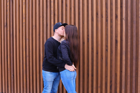 Beautiful young lovers talking about abstract topics, gently hug and talk, kiss, met after long separation, spend weekend together and stand in background of wall outdoors. Man dressed in black sweater, blue jeans, sneakers and black cap, girl with long bの写真素材