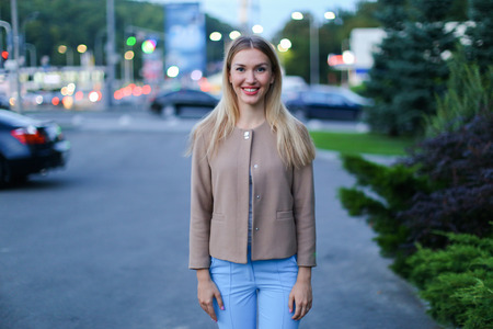 Women with blond hair european appearance smiles and removed advertising for clothes and accessories, womens magazine or waiting for friend. Girl dressed in bright light blouse, beige jacket and blue trousers. Concept of stylish girl, shooting advertisingの写真素材