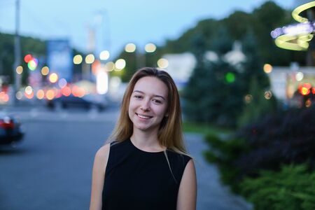Young cute girl with light brown hair smiles broadly at camera, removed for advertising of cosmetics or clothing catalog, awaiting meeting with friend and stands against background of glowing lights urban roads outdoors. Girl dressed in black T-shirt and の写真素材