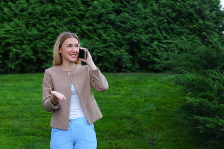 Young modern woman chatters with friend on smartphone, tells funny story and jokes, listens attentively interlocutor at other end of line, shares secrets, shows emotion of joy and gestures. Blonde with long hair dressed in light blouse, beige jacket and bの写真素材