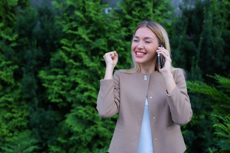 Young modern woman chatters with friend on smartphone, tells funny story and jokes, listens attentively interlocutor at other end of line, shares secrets, shows emotion of joy and gestures. Blonde with long hair dressed in light blouse, beige jacket and bの写真素材