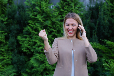 Young modern woman chatters with friend on smartphone, tells funny story and jokes, listens attentively interlocutor at other end of line, shares secrets, shows emotion of joy and gestures. Blonde with long hair dressed in light blouse, beige jacket and bの写真素材