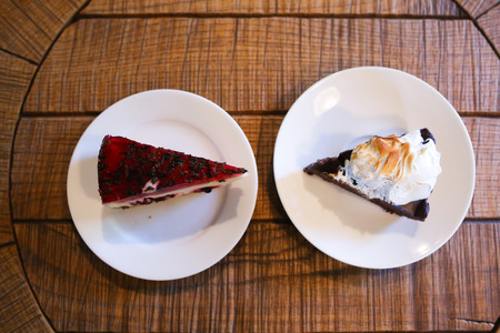 Top view of piece of cherry cheesecake with jelly and chocolate cake decorated with whipped cream on white round plates, which stand on round wooden table in cafe. Concept of delicious food, pleasure, confectionery, restaurant business.の写真素材