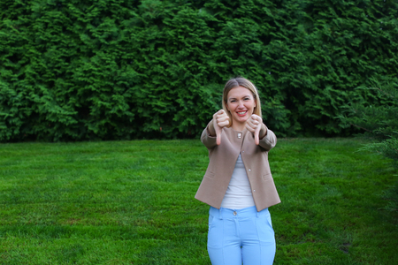 Slender blonde curves and shows gesture "Bad" grieved and shows evaluation of product, does not recommend fitness club or restaurant. Women with long blonde hair dressed in bright blouse, beige jacket and blue pants, standing on background of green grass の写真素材