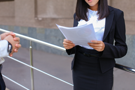 Pretty female girl student holds in hands important paper, Students work or homework, constitutes plan and stands against backdrop of modern business center.Woman with black hair, dressed in white blouse and classic jacket with black skirt and heels.Conceの写真素材