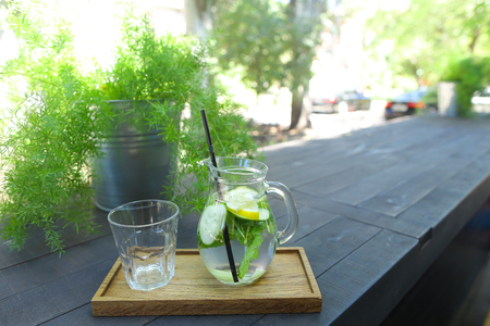 Glass decanter and black drinking straw with refreshing drink from cucumber, lemon, mint and empty glass faceted cup stands on wooden board on gray wooden table on background of green flowers in gray bucket, iron pot in cafe. Concept of refreshing drink, の写真素材