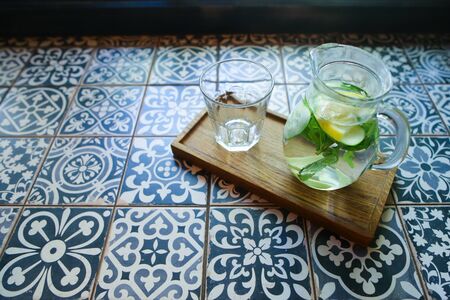 Glass decanter with refreshing drink from cucumber, lemon, mint and empty glass faceted cup stands on tile with patterns in cafe. Concept of refreshing drink, non-alcoholic cocktails, restaurants business, menu of beverages.の写真素材