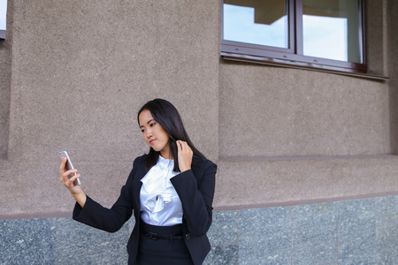Charming cute lady girl student photographed, makes selfie photos and smiling at camera on background of wall business building outdoors. Woman with black hair, dressed in white blouse, classic jacket and black skirt with heels .Concept of takes selfie, cの写真素材