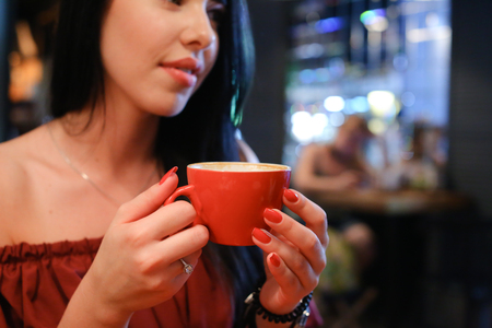 Lovely young female girl holds in hand red cup and drinks coffee behind wooden square table against background of interior and people in cozy coffee shop with gray modern lamp. On table stand desserts and holder for napkins. Girl with black hair dressed iの写真素材