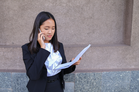 Serious young adult business woman  talks on phone, solves problems, holds and looks at documents and stands on background of gray wall of business center. Woman with black hair, dressed in white blouse and classic jacket with black skirt and heels. Conceの写真素材