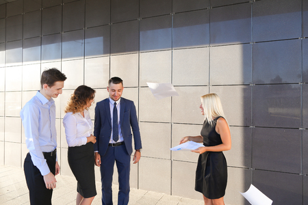 Beautiful young ambitious girl in black dress holds paper and explains, makes remarks, explains adjustments in documents, share information and advice with colleagues, who listen, contribute their ideas and stand on wall background of business center outdの写真素材