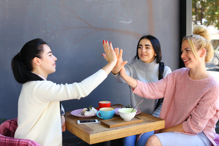 Three young attractive girls girlfriends rejoice, high five to each other, chatter, gossiping, share secrets, laughing, smiling and sitting on chair at table on background of wall and trees in coffee shop. Girls with black hair dressed in  white and gray の写真素材