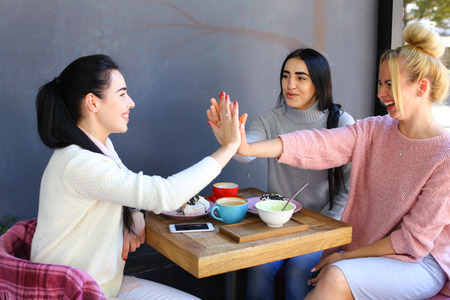Three young attractive girls girlfriends rejoice, high five to each other, chatter, gossiping, share secrets, laughing, smiling and sitting on chair at table on background of wall and trees in coffee shop. Girls with black hair dressed in  white and gray の写真素材