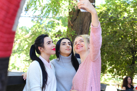 Three wonderful young cute female girl girlfriends make selfie, photo on phone by heart, smiling and laughing in cafe on background trees outdoors. Girls with black hair dressed in white and gray knitted sweaters , girl with blonde hair dressed in pink knの写真素材