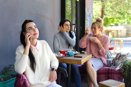 Three young magnificent girl girlfriends relaxing in cafÃ©. One of girls speaks by phone and smile on background of girlfriends, who laugh, communicate, share secrets, gossiping and eat at table in cafe. Girls with black hair dressed in  white and gray knの写真素材