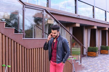 Confident Attractive Young Guy Muslim Businessman Goes Slowly Along Wall, Holding Phone in Hand and Talks, Resolve Issues, Discuss Future Plans and Smiling, Holds Hand in Pocket, Stands on Background of Wooden Panel Stairs Near House Outdoors. Bearded Manの写真素材