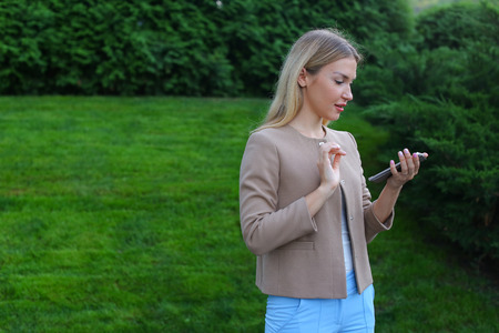 Pretty blonde european appearance uses smartphone for correspondence with friends, make an appointment, looks at screen of gadget, select gifts for friends and viewing news feed. Girl dressed in light blouse, beige jacket and blue pants and stands in profの写真素材