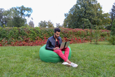 Attractive Young Arabic Guy, Businessman, Student Sits With Laptop in Soft Green Chair, Looks at Computer Screen and in Aside, Speculates About Work, Thinking About Solving Problems, Scratches Beard and Smiling, Working Online, Remotely on Background of Gの写真素材