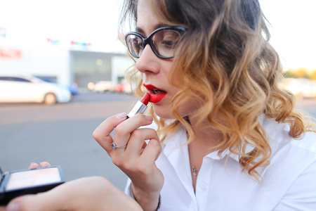 Young beautiful confident woman holds in hands compact powder, looking in mirror and paints lips red lipstick on street. Girl with curly hair and in glasses dressed in white blouse. Concept of make up, cosmetics, lunch break, confident business woman.の写真素材