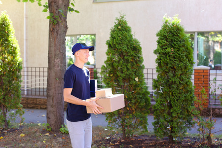 Deliveryman Carring Parcel and Giving it and Smiling. International Staffing Company Shows That Courier of Company Always Ready to Work at Any Time and Any Day, Young Man of European Appearance in Uniform, With Broad Smile and Good Mood on Background of Hの写真素材