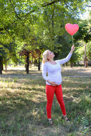 Alluring Future Mother of European Appearance Enjoys Pleasant Things and Look Forward to Appearance of First-Born Into World Photographed For Advertising of Womens Clothing For Pregnant With Pink Ballon in Shape of Heart in Green Sunny Park Outdoors in Daの写真素材