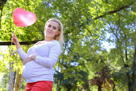 Marvelous Young Adult Lady With Child of European Appearance Enjoying Sunny Day, Cute Smiles, Poses and Rises up Pink Ballons in Shape of Heart in Green Park Outdoors in Daytime. Female With Blonde Hair Braided in Braid in Front Dressed in White Blouse Wiの写真素材
